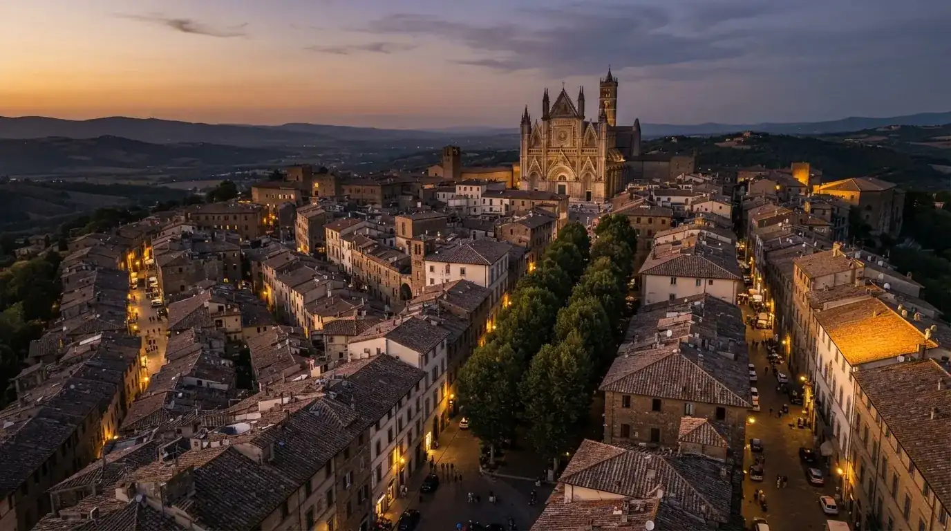 Vista dall'alto di una città italiana al tramonto, con luci che si accendono nelle finestre degli edifici lungo una via trafficata.